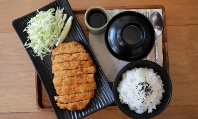 Crispy tonkatsu with shredded cabbage and a lemon wedge on a black plate, served with white rice (sesame seeds) and dipping sauce on a wooden table.
