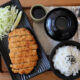 Crispy tonkatsu with shredded cabbage and a lemon wedge on a black plate, served with white rice (sesame seeds) and dipping sauce on a wooden table.