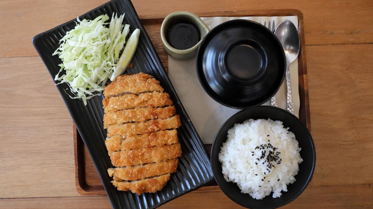 Crispy tonkatsu with shredded cabbage and a lemon wedge on a black plate, served with white rice (sesame seeds) and dipping sauce on a wooden table.