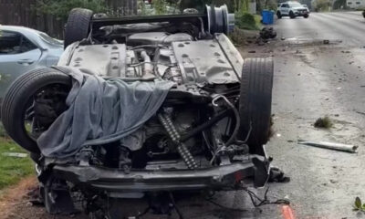 Overturned car on a residential street with severe front-end damage; debris scattered and a gray cloth draped over the engine area.
