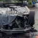 Overturned car on a residential street with severe front-end damage; debris scattered and a gray cloth draped over the engine area.