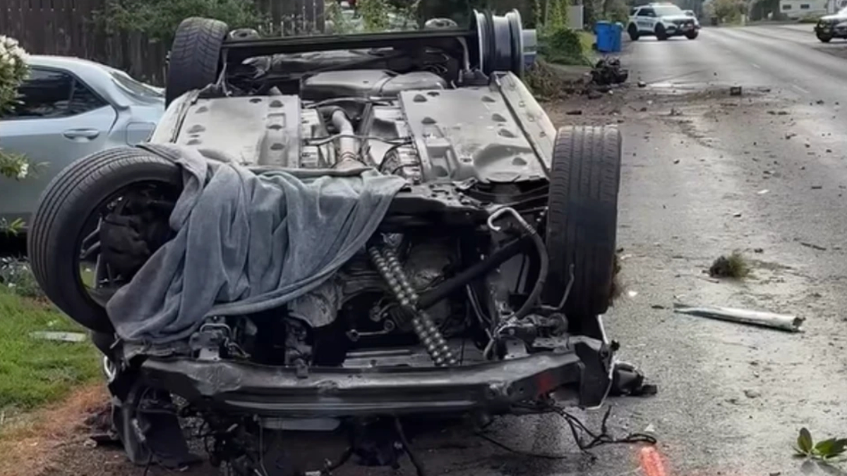 Overturned car on a residential street with severe front-end damage; debris scattered and a gray cloth draped over the engine area.