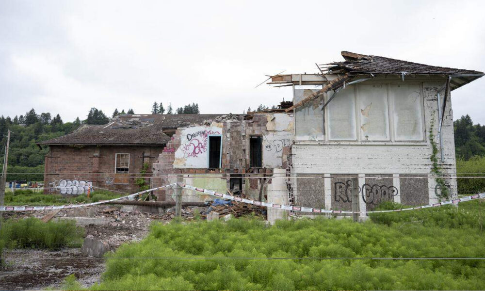 Abandoned, partially collapsed brick building with graffiti, surrounded by caution tape in a grassy lot.