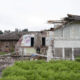 Abandoned, partially collapsed brick building with graffiti, surrounded by caution tape in a grassy lot.