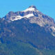 Snow-capped mountain peak rising above dense evergreen forest under a clear blue sky.