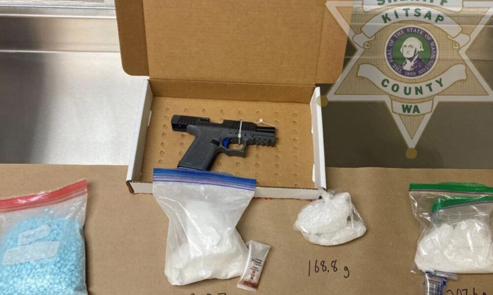 Semi-automatic pistol in an open cardboard evidence box, surrounded by bags of white powder and a small vial, with a handwritten weight (168.9 g) on the table and a sheriff emblem in the background.