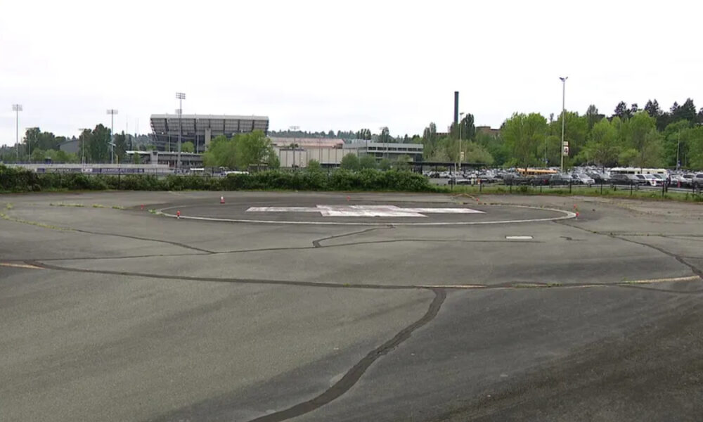 Empty circular helicopter landing pad marked with an H in a large paved lot, with a stadium and trees in the background.