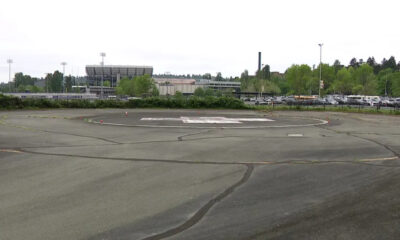 Empty circular helicopter landing pad marked with an H in a large paved lot, with a stadium and trees in the background.