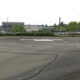 Empty circular helicopter landing pad marked with an H in a large paved lot, with a stadium and trees in the background.