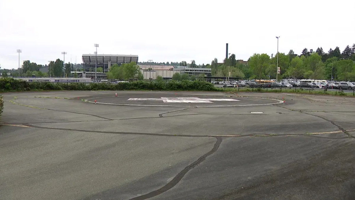 Empty circular helicopter landing pad marked with an H in a large paved lot, with a stadium and trees in the background.