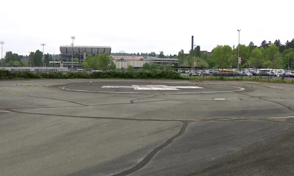 Circular helicopter landing pad ('H') in a large paved lot with trees, a stadium, and a parking lot in the background.
