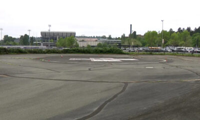 Circular helicopter landing pad ('H') in a large paved lot with trees, a stadium, and a parking lot in the background.