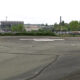 Circular helicopter landing pad ('H') in a large paved lot with trees, a stadium, and a parking lot in the background.