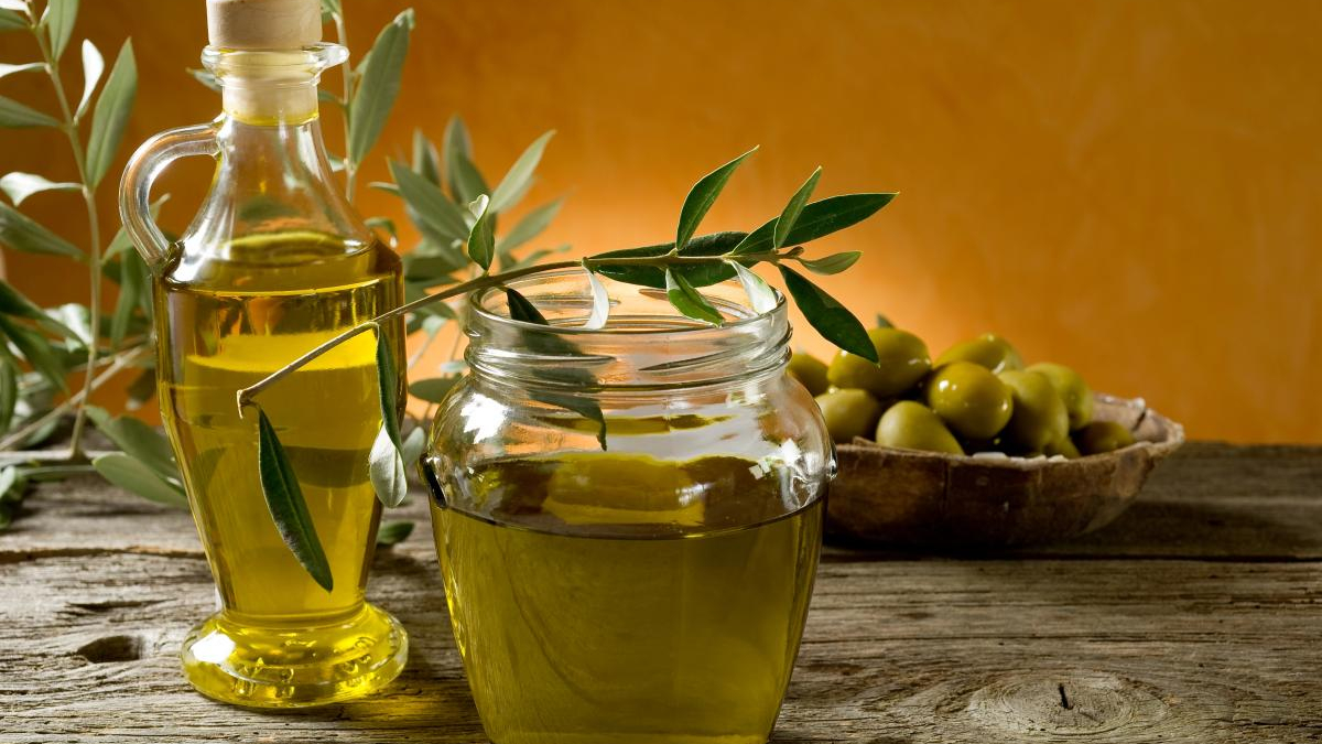 Olive oil bottle and mason jar on a rustic wooden table with olive branches and a bowl of green olives nearby.