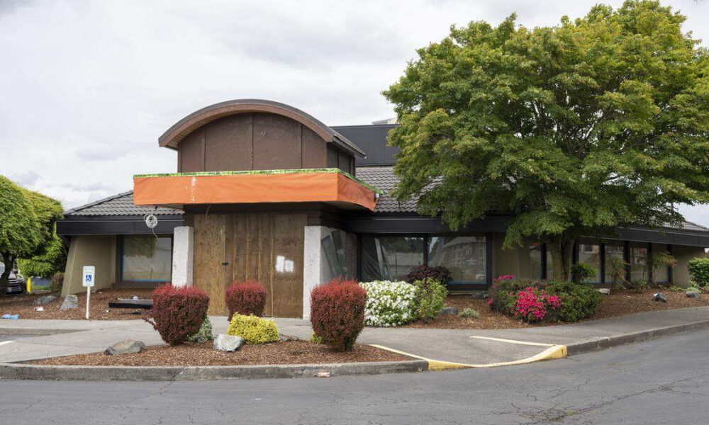 Two-story commercial building with an orange construction awning over a boarded entry, surrounded by shrubs and a large tree in a parking lot.