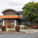 Two-story commercial building with an orange construction awning over a boarded entry, surrounded by shrubs and a large tree in a parking lot.