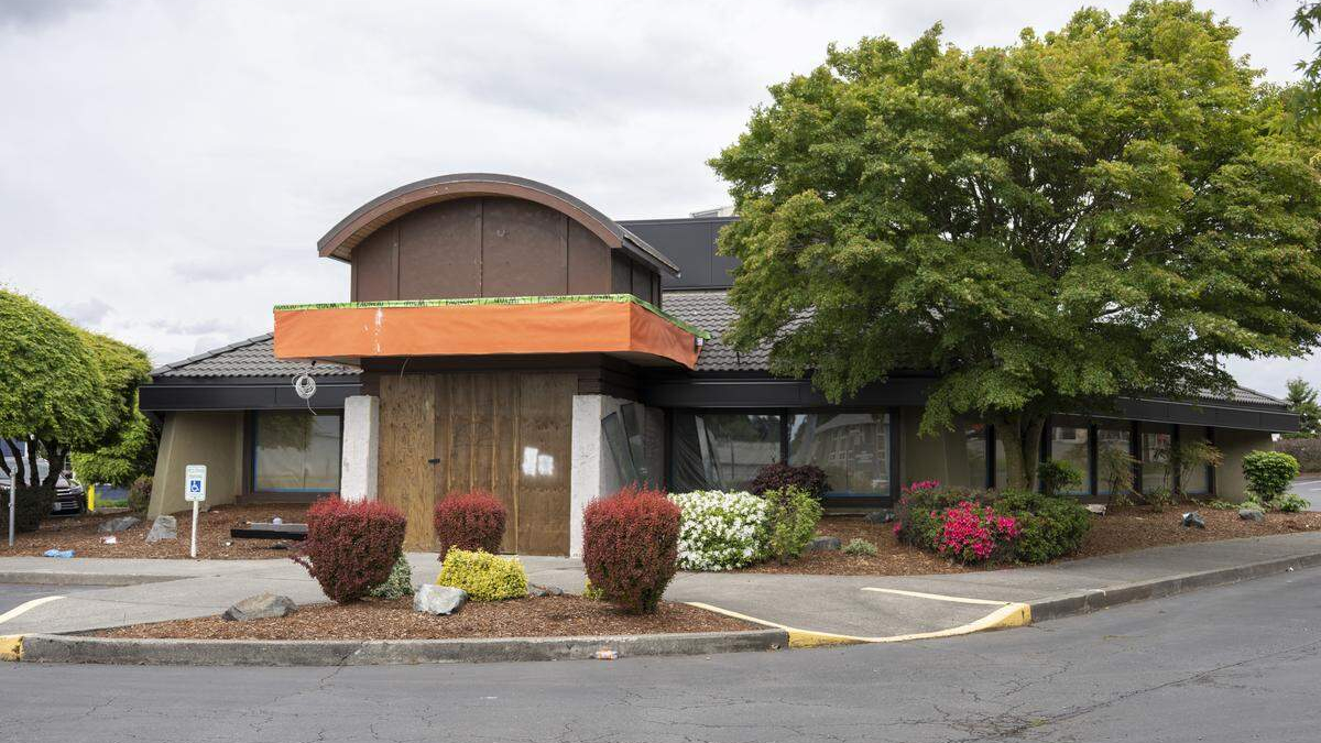 Two-story commercial building with an orange construction awning over a boarded entry, surrounded by shrubs and a large tree in a parking lot.