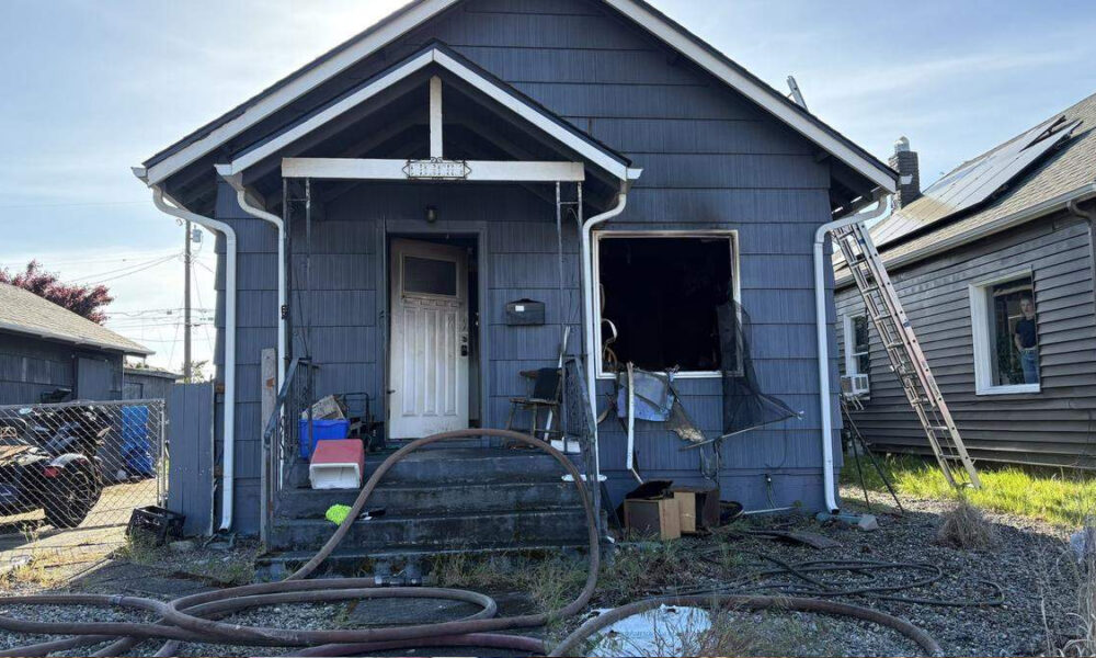 Blue house with a fire-damaged front entrance, broken window, and scattered debris; hoses lie across the yard.