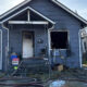 Blue house with a fire-damaged front entrance, broken window, and scattered debris; hoses lie across the yard.