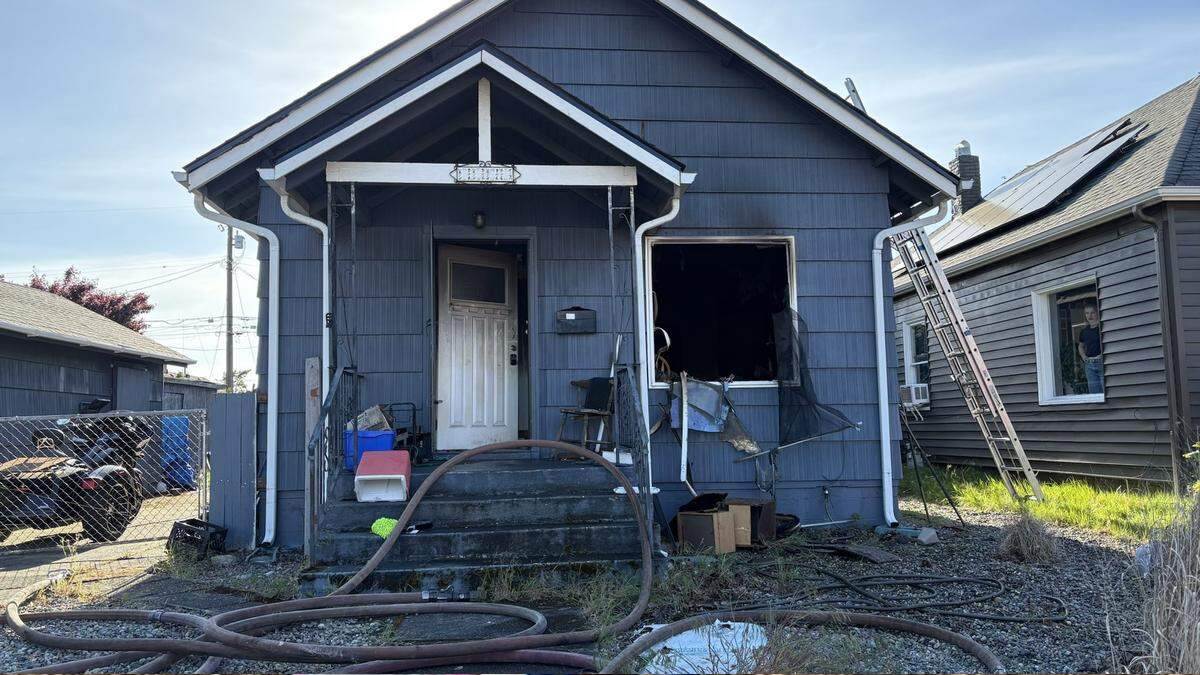 Blue house with a fire-damaged front entrance, broken window, and scattered debris; hoses lie across the yard.