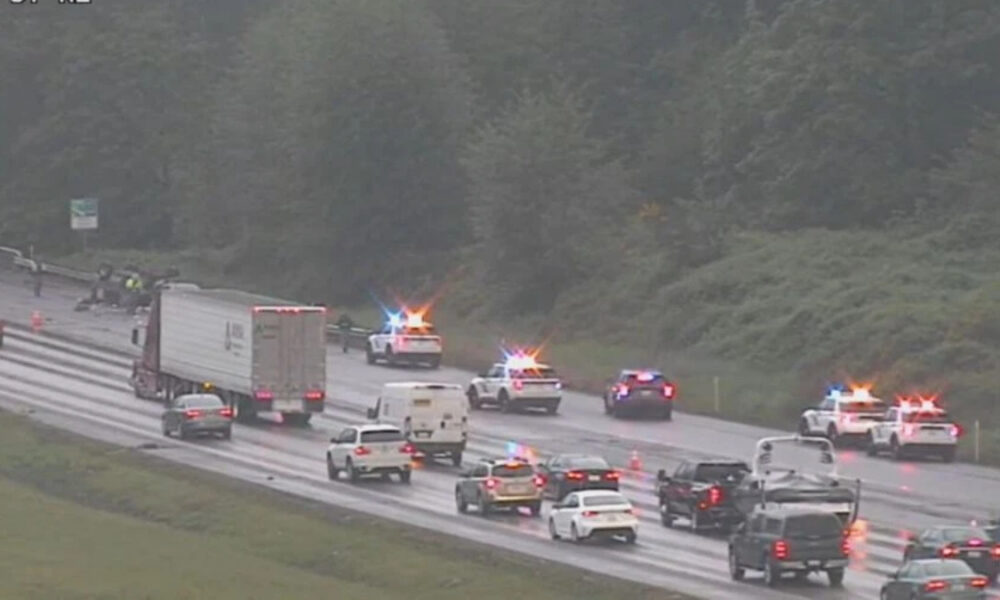 Emergency responders with flashing lights at a multi-vehicle crash on a wet highway, with traffic backed up nearby.