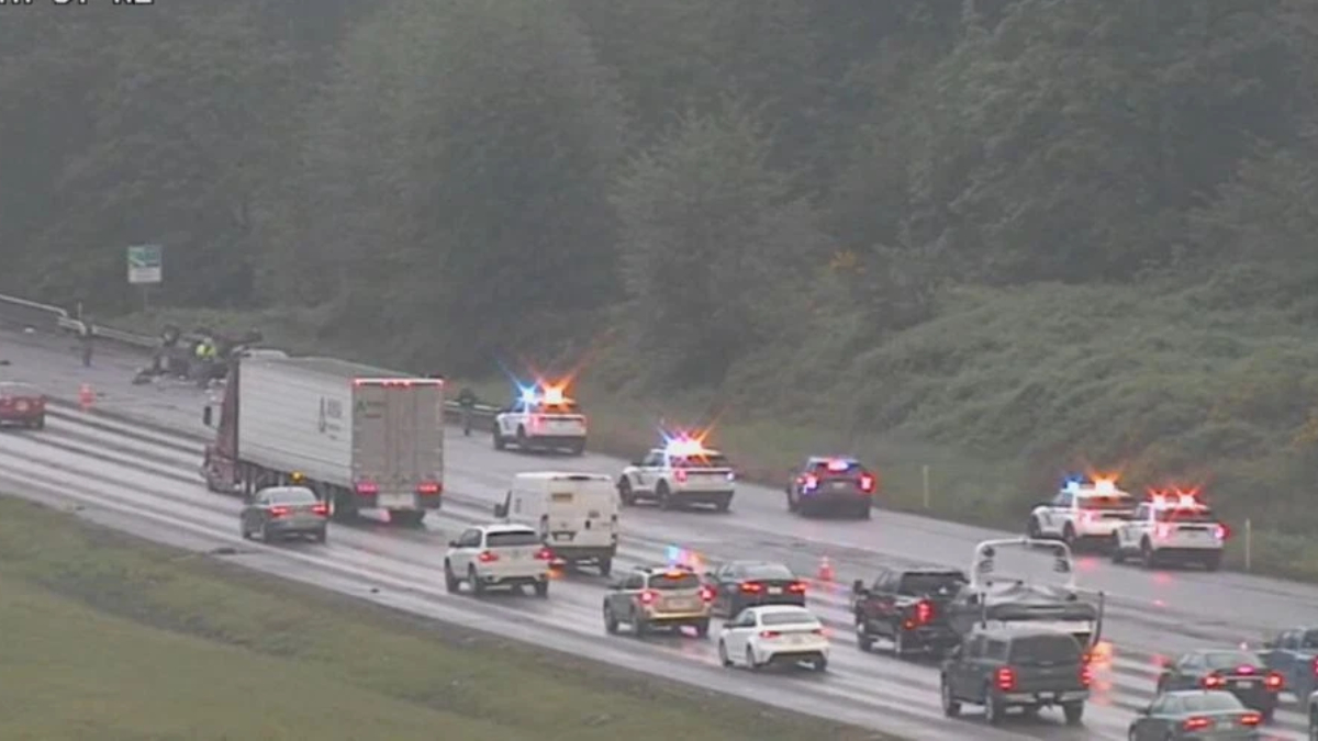 Emergency responders with flashing lights at a multi-vehicle crash on a wet highway, with traffic backed up nearby.