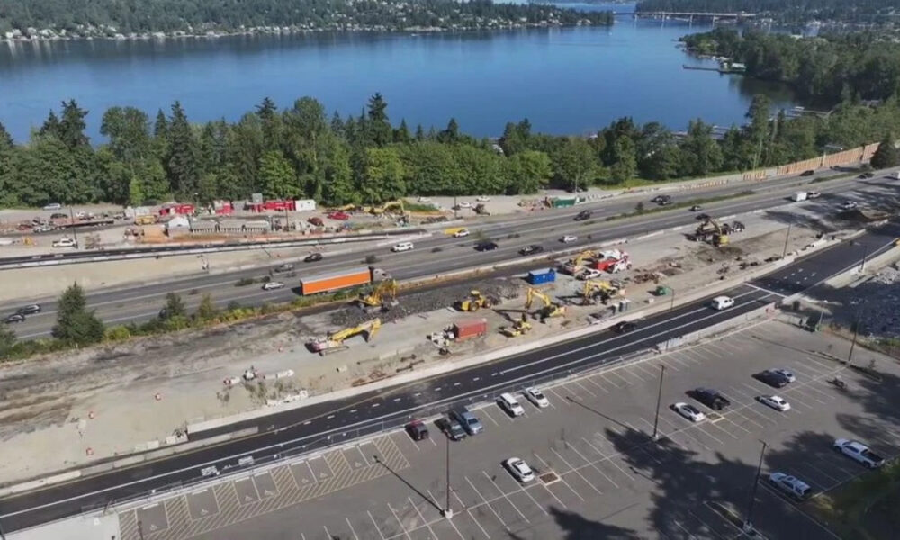 A busy construction site beside a multi-lane highway next to a blue lake, with excavators, trucks, and barriers.