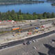 A busy construction site beside a multi-lane highway next to a blue lake, with excavators, trucks, and barriers.