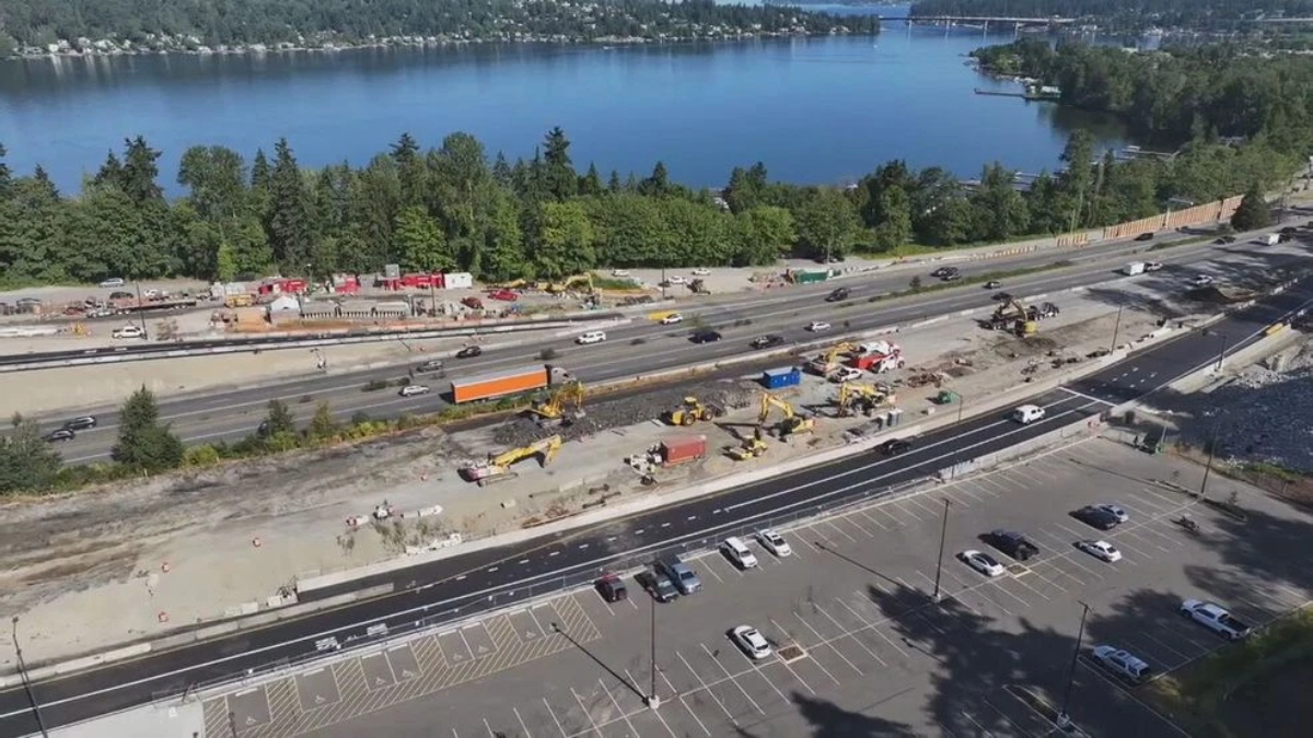A busy construction site beside a multi-lane highway next to a blue lake, with excavators, trucks, and barriers.