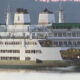 Large white passenger ferry with multiple decks sailing on calm water, rear vehicle loading ramp open for cars.