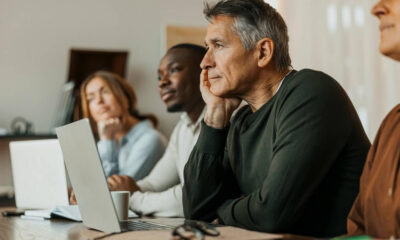 Group of professionals in a meeting room listening intently, with a gray-haired man in a dark sweater resting his chin on his hand in the foreground.