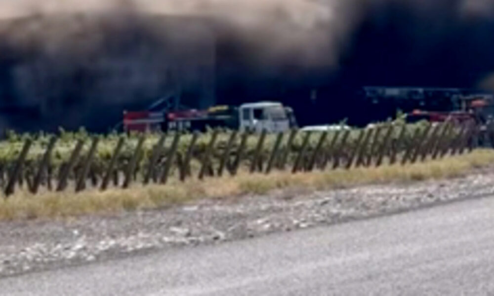 Emergency vehicles and a white van near a smoke-filled tunnel, seen from the roadside with a wooden fence in the foreground