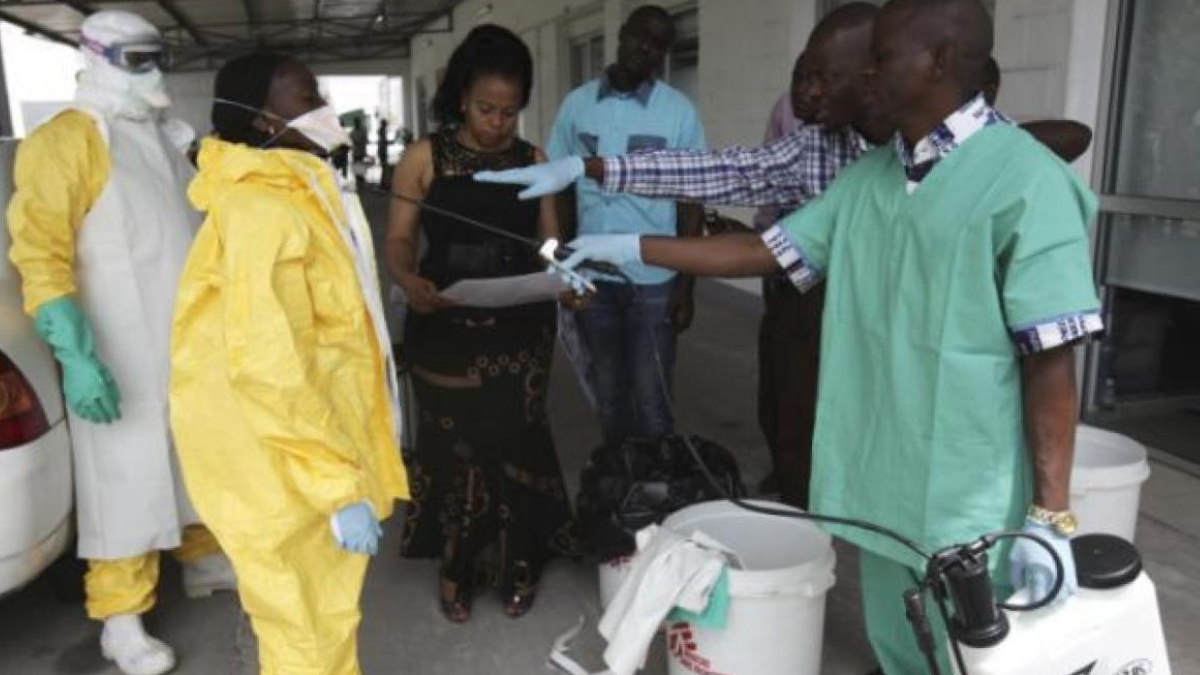 Medical team in PPE (yellow hazmat suit and protective gear) outside, exchanging a document with a woman as they prepare equipment.