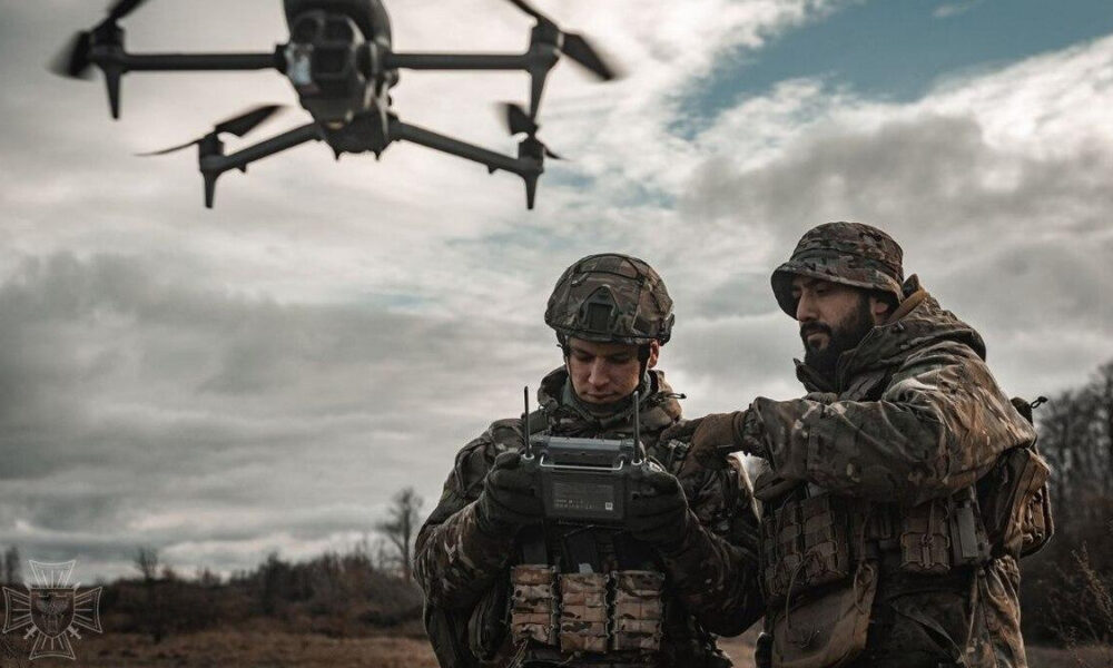 Two soldiers in camouflage gear operate a handheld drone controller as a quadcopter hovers above them in an open field under a cloudy sky.