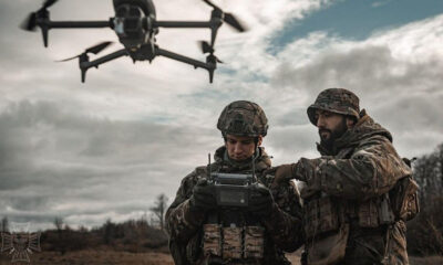 Two soldiers in camouflage gear operate a handheld drone controller as a quadcopter hovers above them in an open field under a cloudy sky.