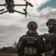 Two soldiers in camouflage gear operate a handheld drone controller as a quadcopter hovers above them in an open field under a cloudy sky.