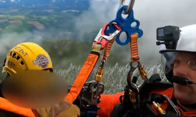 Two rescue workers in orange gear secure a person with ropes and carabiners on a cliff edge; a GoPro camera is mounted on a white helmet nearby.