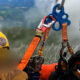 Two rescue workers in orange gear secure a person with ropes and carabiners on a cliff edge; a GoPro camera is mounted on a white helmet nearby.