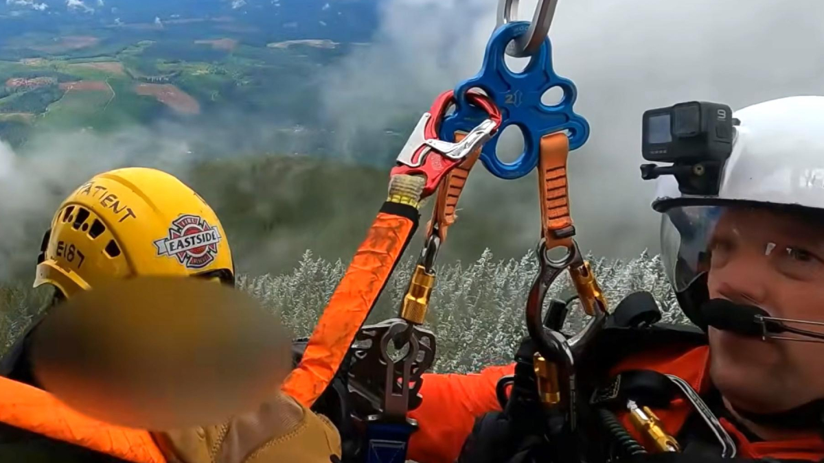 Two rescue workers in orange gear secure a person with ropes and carabiners on a cliff edge; a GoPro camera is mounted on a white helmet nearby.