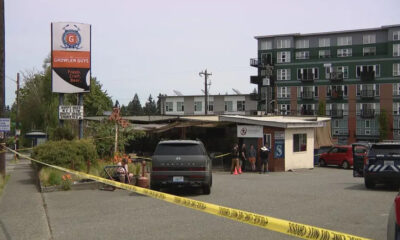 Police tape blocks a parking lot in front of a small storefront as a gray SUV and a few people stand near the entrance, with a green apartment building in the background.