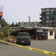 Police tape blocks a parking lot in front of a small storefront as a gray SUV and a few people stand near the entrance, with a green apartment building in the background.