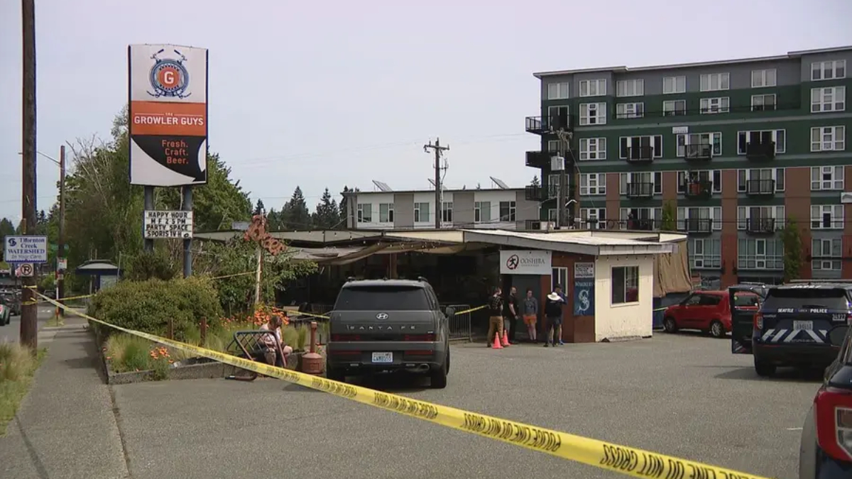 Police tape blocks a parking lot in front of a small storefront as a gray SUV and a few people stand near the entrance, with a green apartment building in the background.