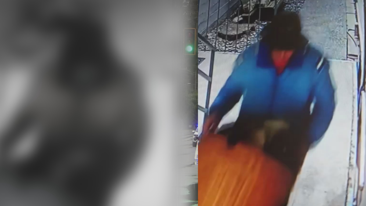 Surveillance still of a person in a blue jacket walking on a sidewalk, captured by a security camera on the right side of the frame.
