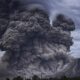 Volcanic eruption with a massive dark ash cloud rising from a mountain, billowing into the sky.