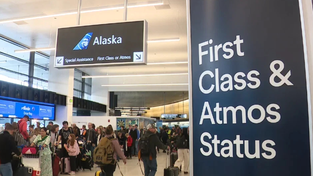 Crowded airport check-in area with travelers and luggage under an Alaska Airlines sign; a tall dark panel on the right displays large white text reading 'First Class & Status' (or 'First Class & Atoms Status').