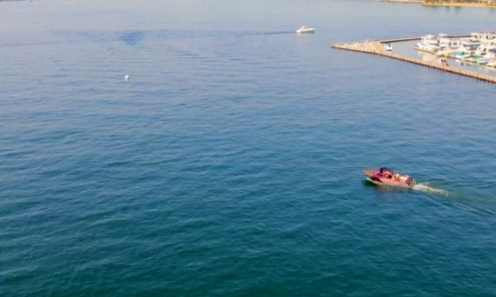 Red and white motorboat with passengers cruising through calm blue water near a marina and breakwater on the right side.