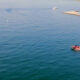 Red and white motorboat with passengers cruising through calm blue water near a marina and breakwater on the right side.