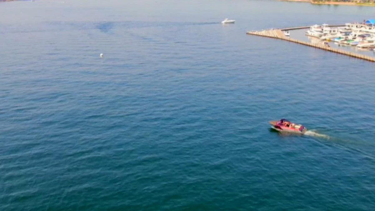 Red and white motorboat with passengers cruising through calm blue water near a marina and breakwater on the right side.