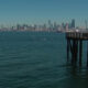 Calm harbor with a distant city skyline and a wooden pier extending from the right into the water, under a clear blue sky.