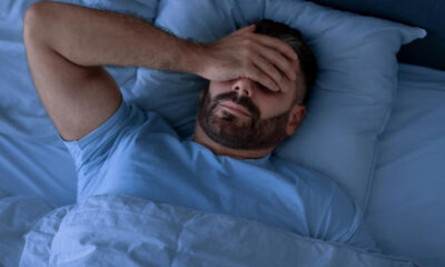 Man with a beard lies in bed under blue bedding, hand covering his eyes in a gesture of fatigue or stress.